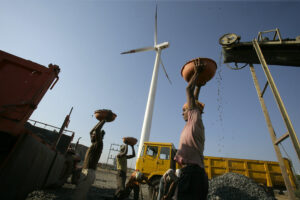 A laborer is seen working at a deisel powered crusher infont of a wind turbine.